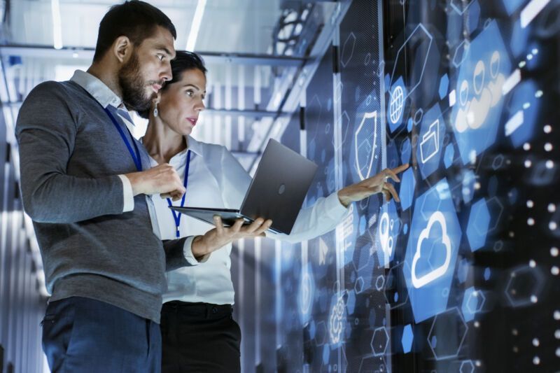 Male IT Specialist Holds Laptop and Discusses Work with Female Server Technician. They're Standing in Data Center, Rack Server Cabinet with Cloud Server Icon and Visualization.